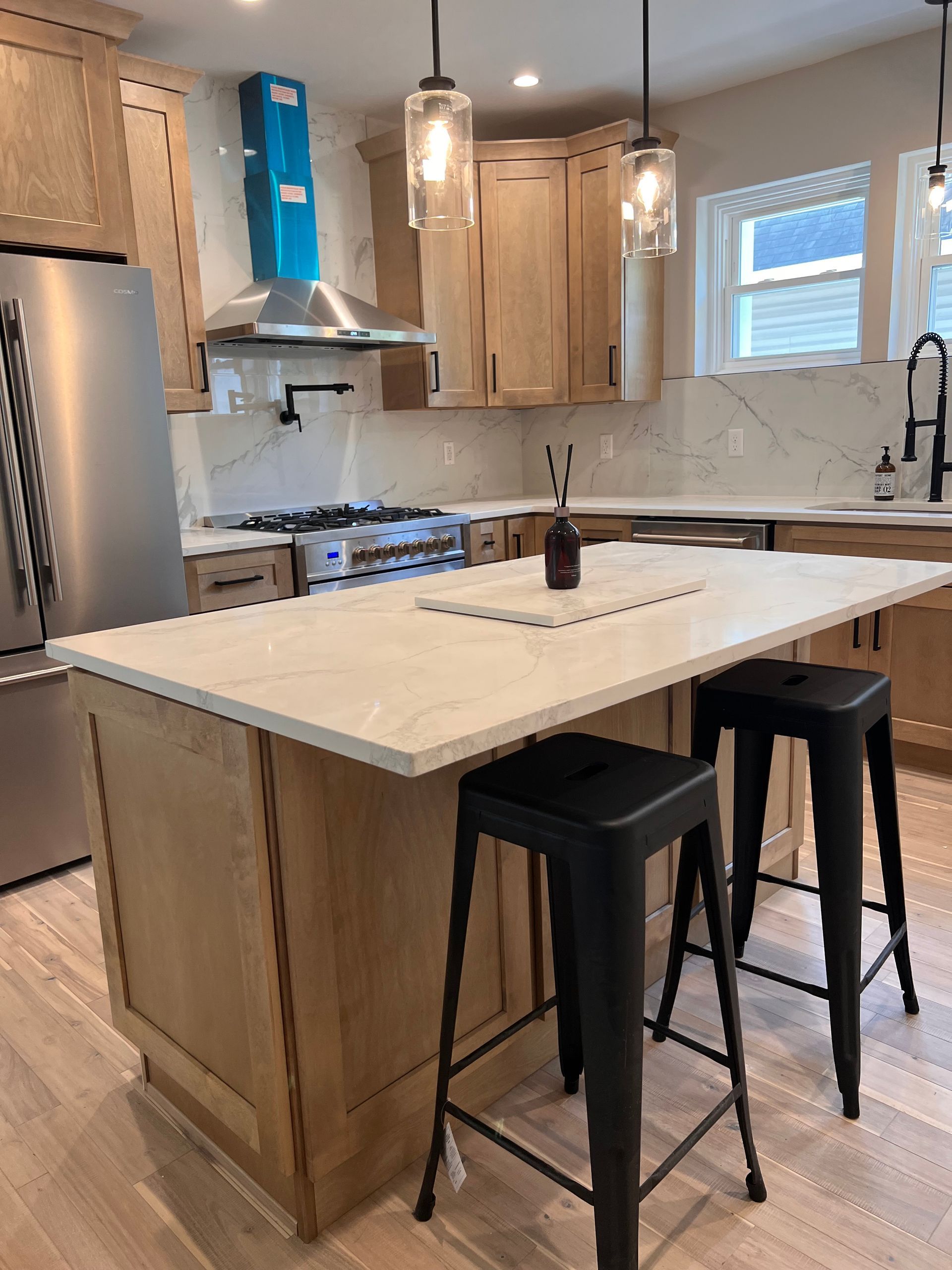 Modern kitchen with light wood cabinets, white countertops, island with two black stools, and stainless steel appliances.