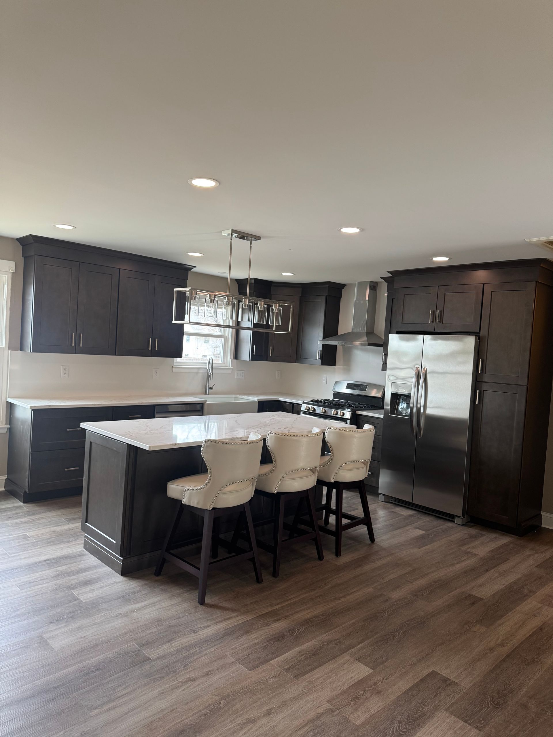 Modern kitchen with dark cabinetry, white countertops, island with bar stools, stainless steel appliances, and wood-look flooring.