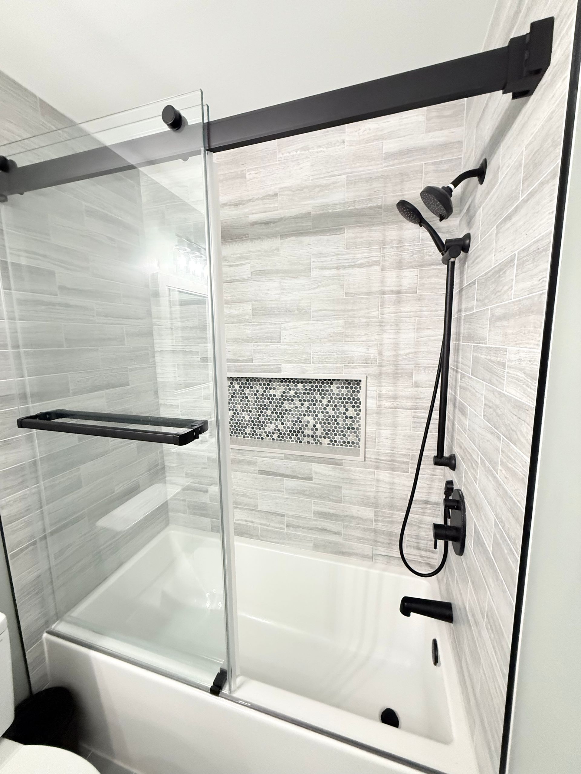 Black-accented shower with sliding glass door, white tub, gray tiled walls, and black shower fixtures.