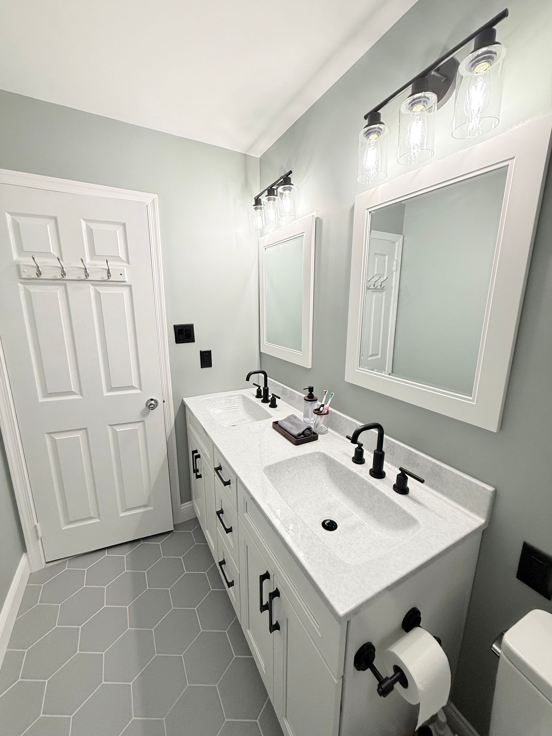 Bathroom with white vanity, black hardware, two sinks, and mirrors. Light green walls, gray tile floor.