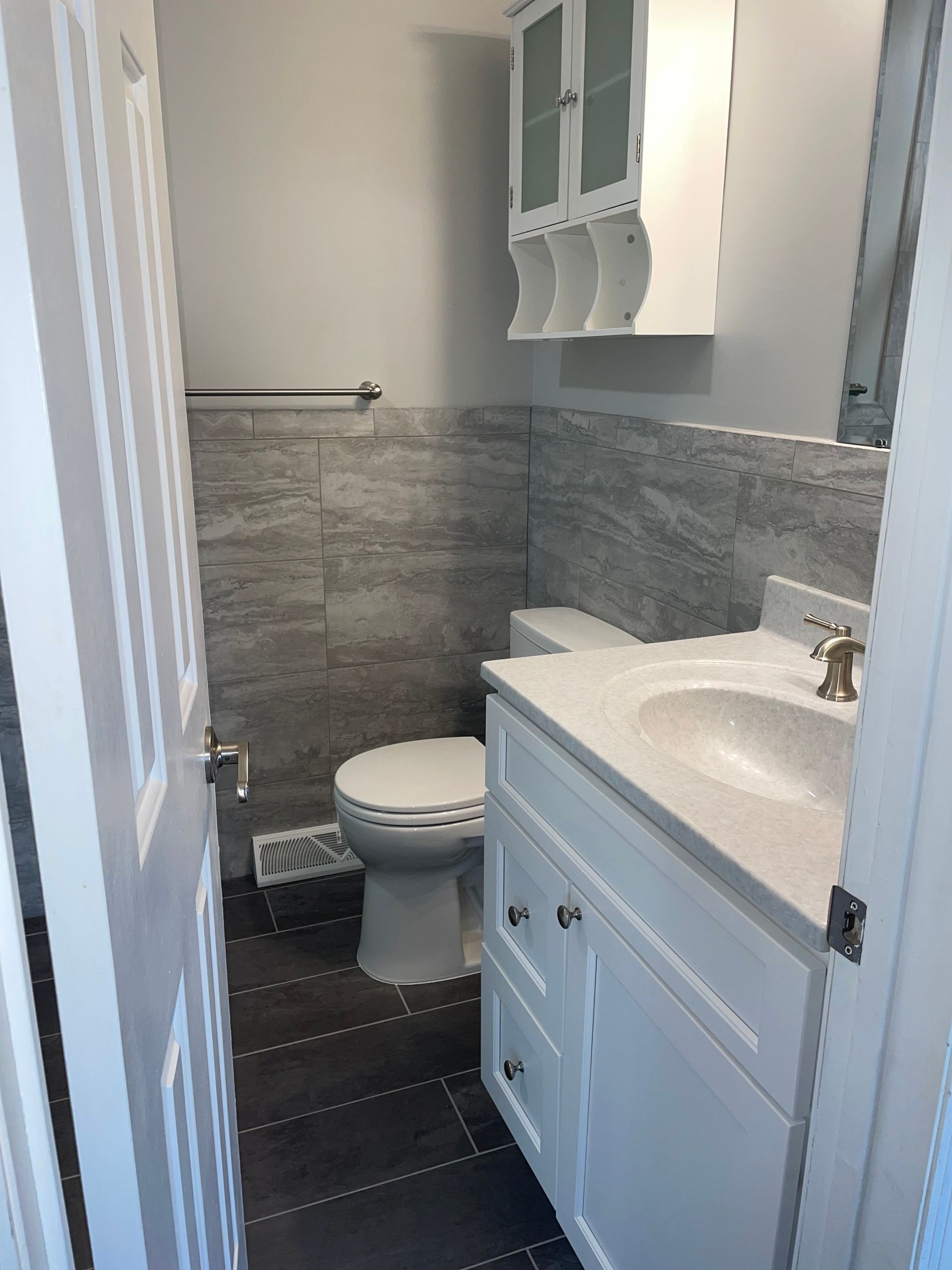 Small white bathroom with toilet, sink, and gray tiled wall. White cabinet above the toilet.