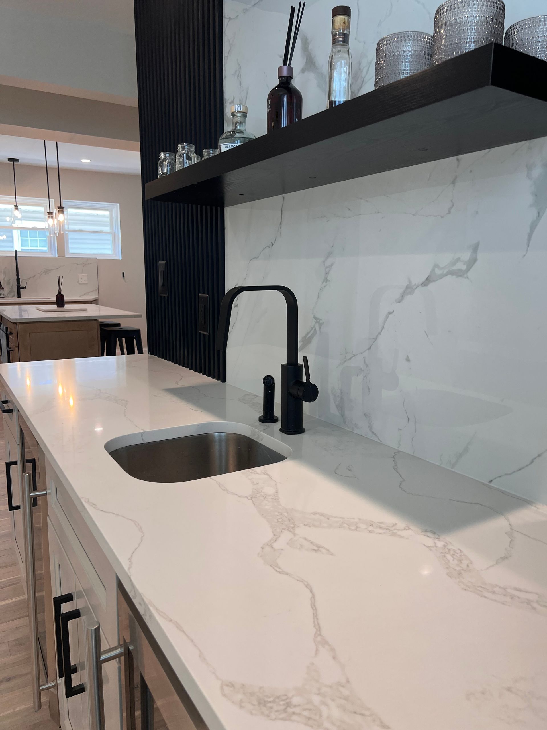Modern kitchen bar with white countertop, black faucet, sink, and floating shelf with glassware.