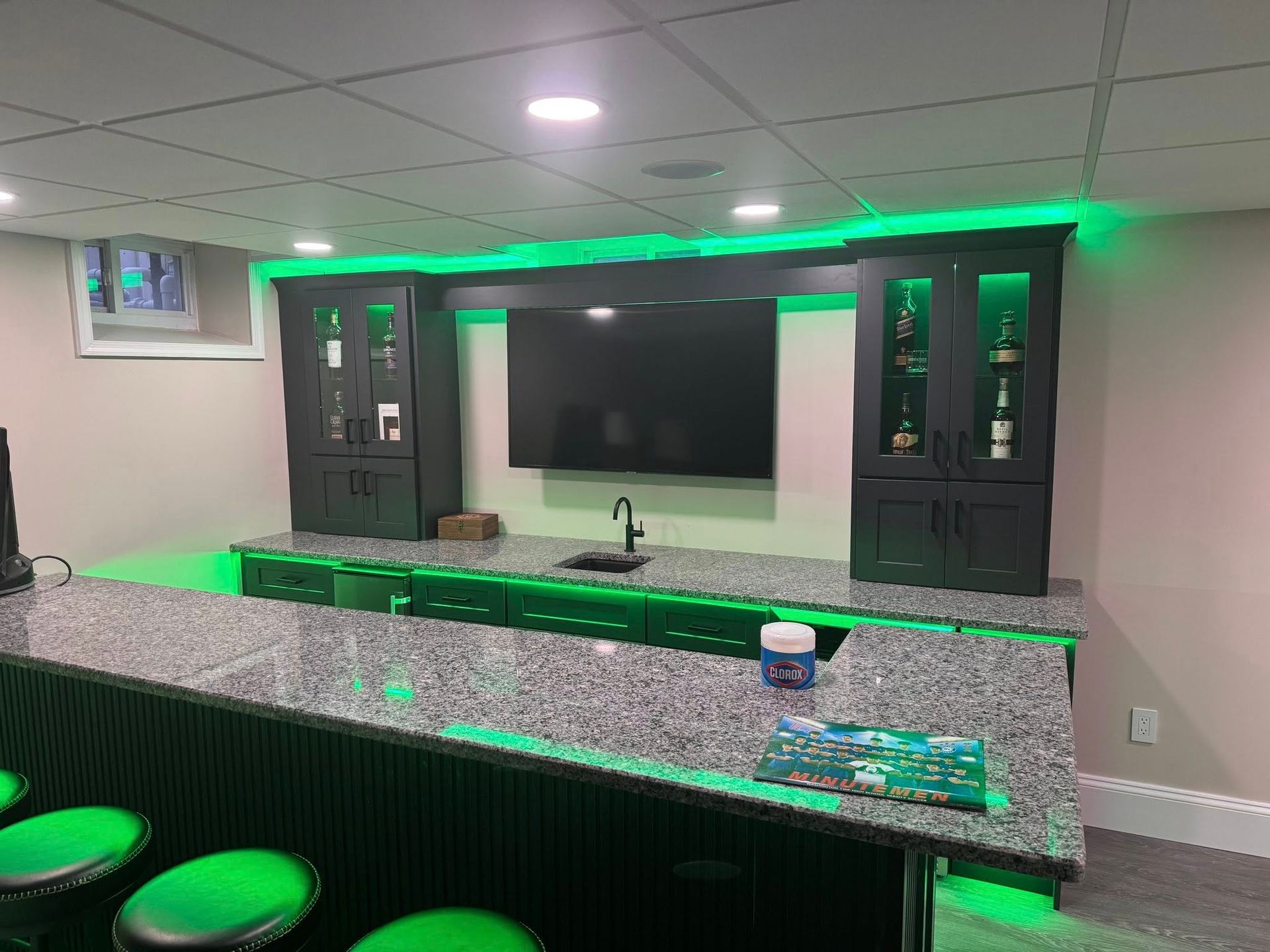 Basement bar with granite countertop, cabinets, sink, TV, and green LED lighting.