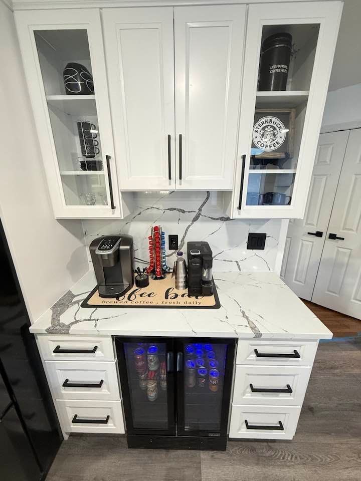 White coffee bar with cabinets, a mini-fridge, and marble countertop; two coffee makers and decor on top.