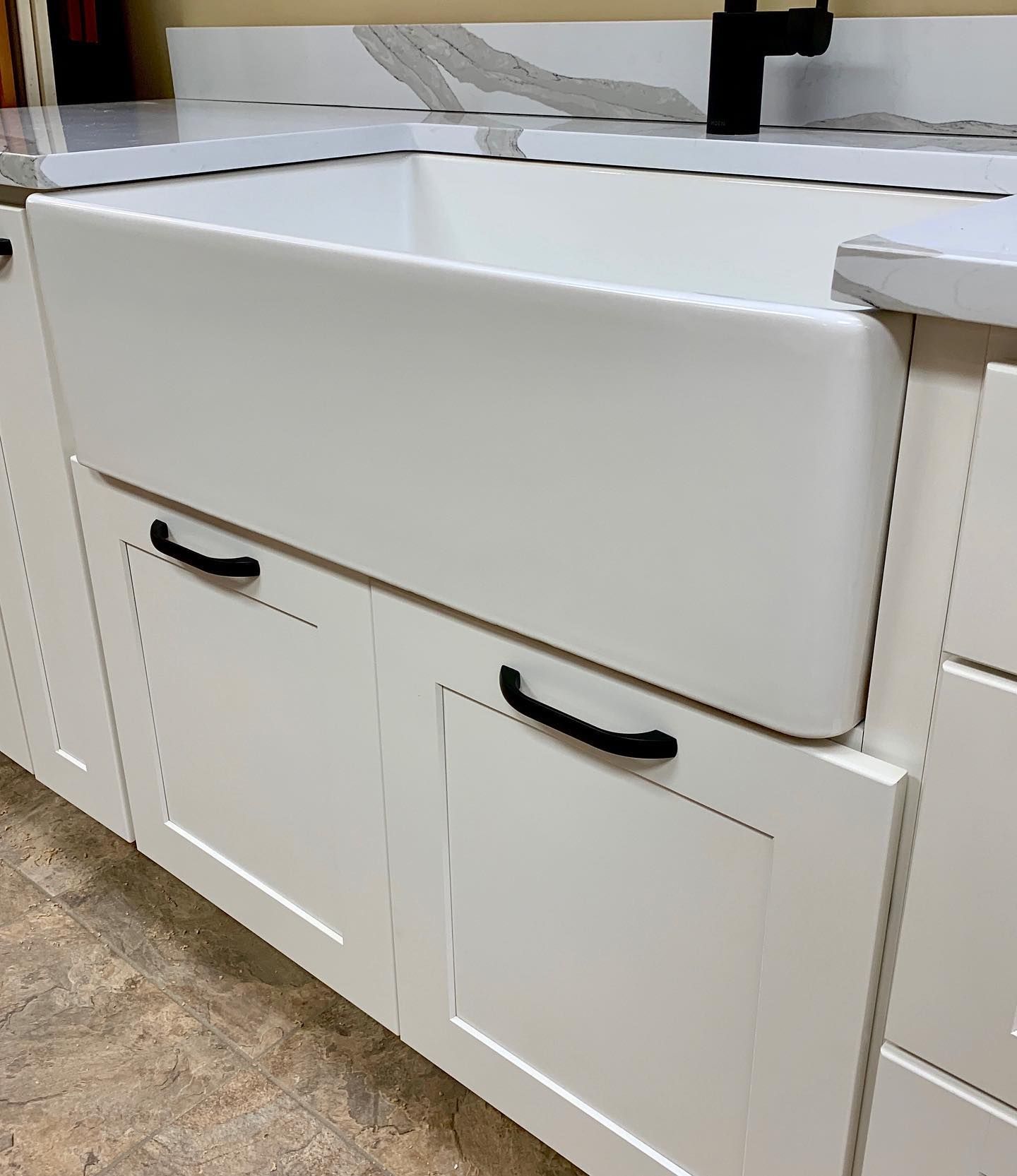 White farmhouse sink in a kitchen cabinet, white and gray countertop, black hardware.