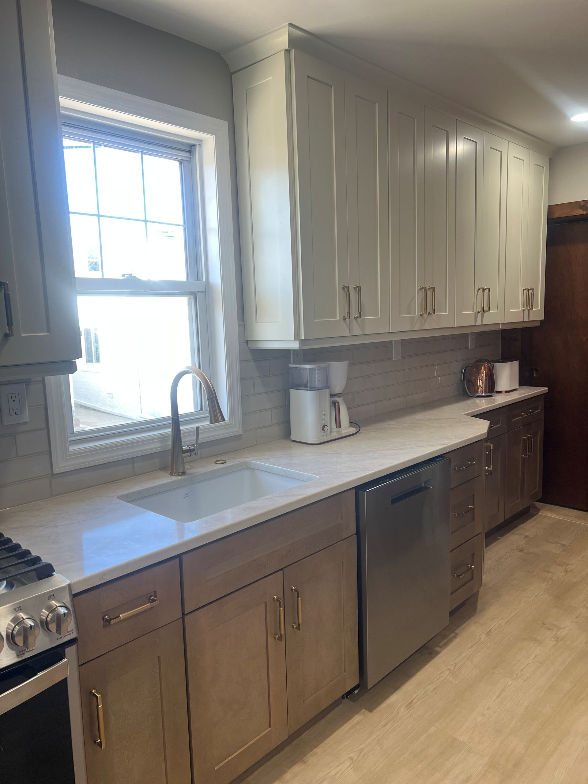 A kitchen with light wood lower cabinets, white upper cabinets, white quartz countertops, a window, and a dishwasher. 