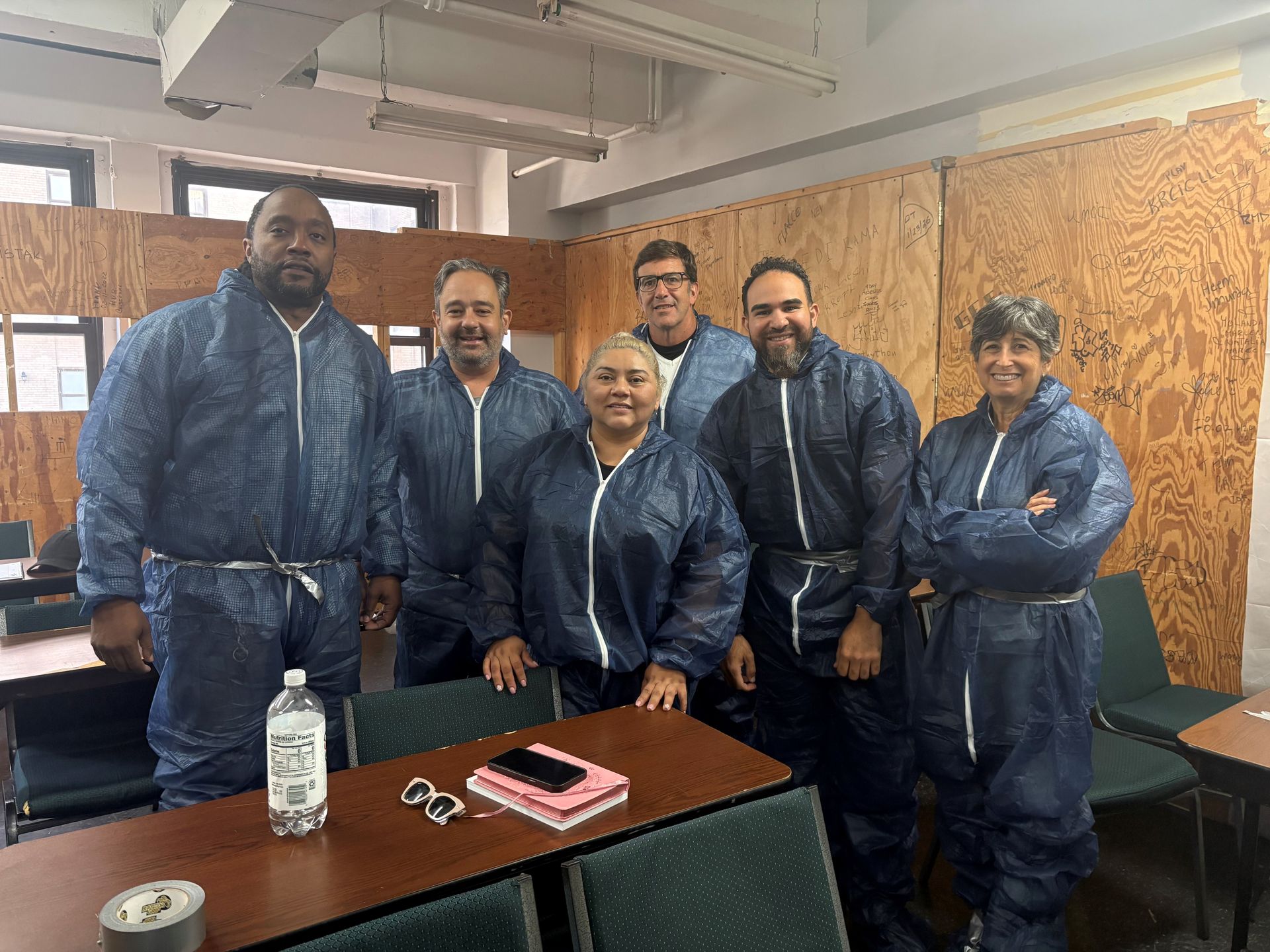 Six people in blue coveralls stand together in a workshop, with a table and wall panels behind them.