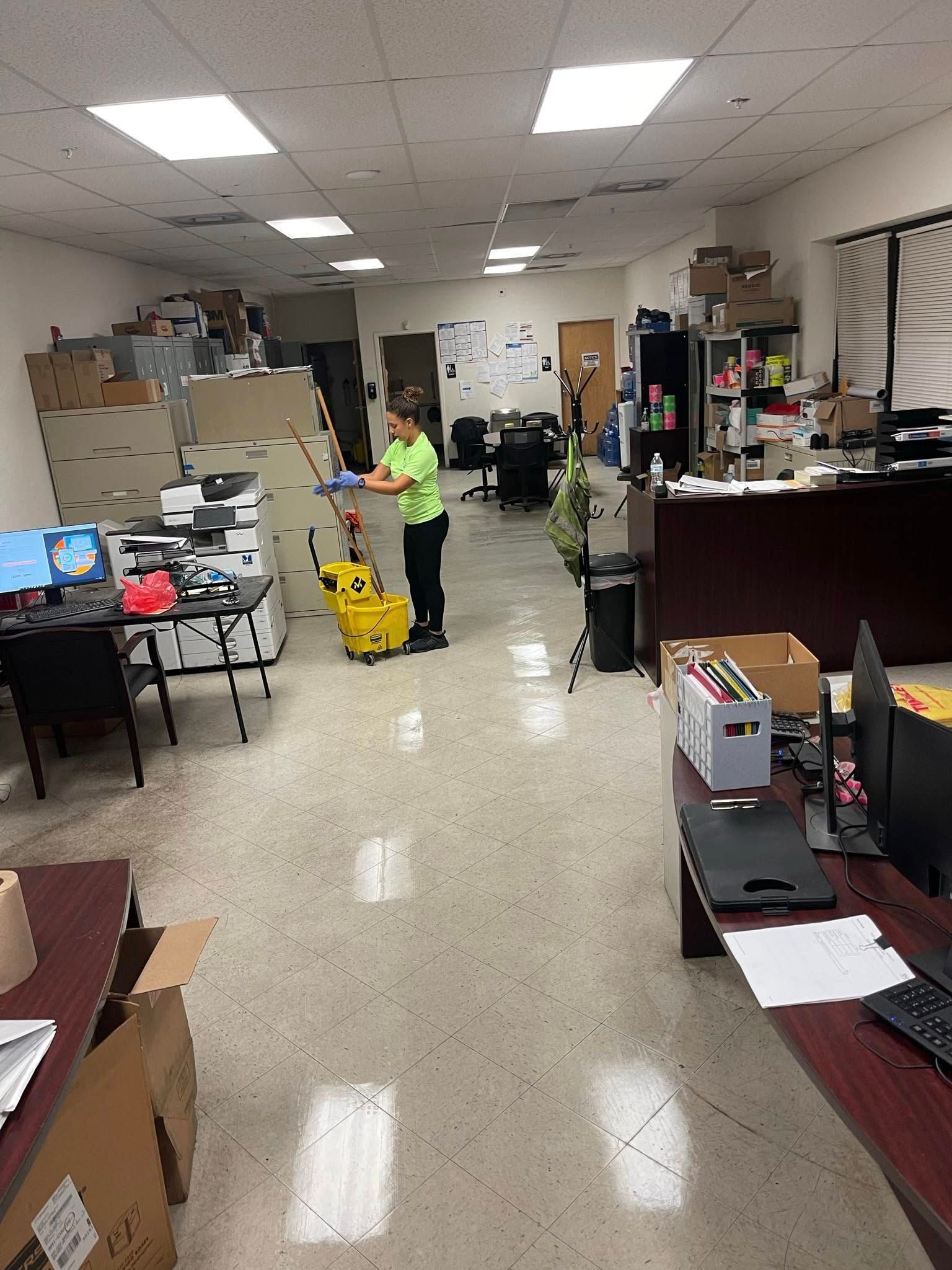Person mopping an office floor; beige and gray decor, cluttered desks, filing cabinets, and overhead lighting.