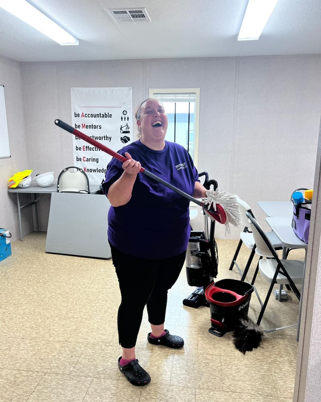 Woman in purple shirt laughing, holding a mop in an office-like setting.