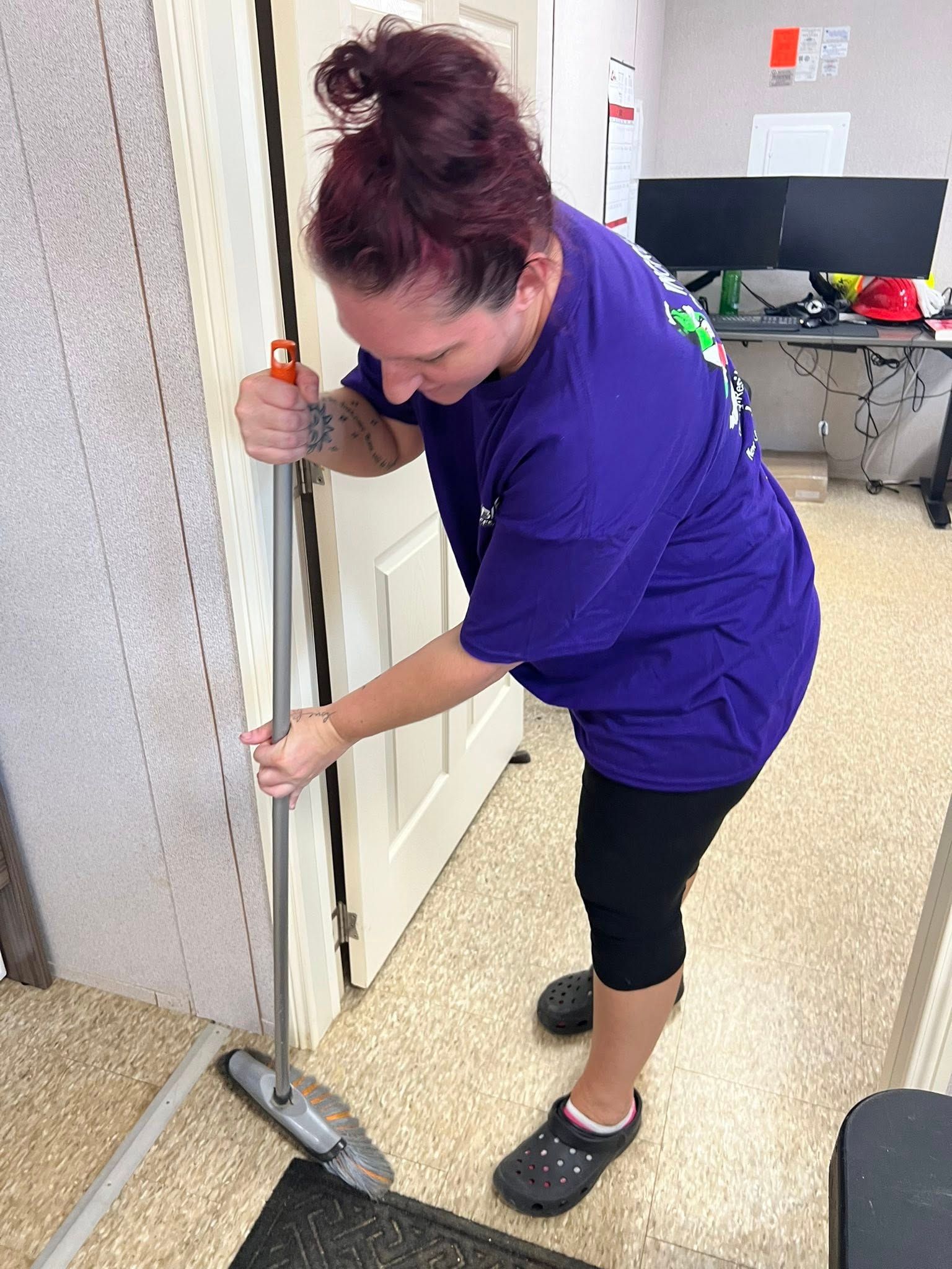 Woman in purple shirt cleans floor with a long-handled mop near a door and office space.