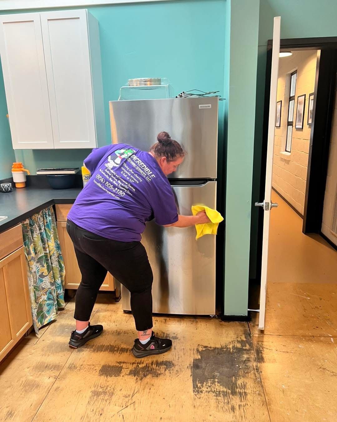 Woman in purple shirt cleaning a stainless steel refrigerator in a kitchen with turquoise walls and light wood cabinets.