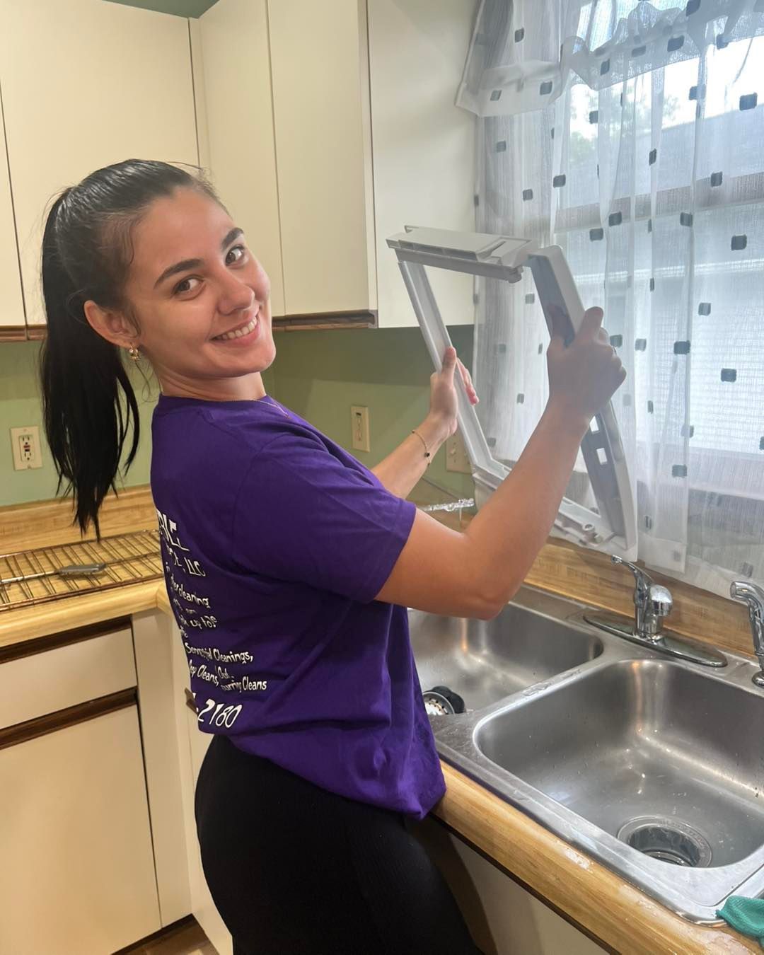 Woman in purple shirt smiles while cleaning a window frame in a kitchen, sink visible.
