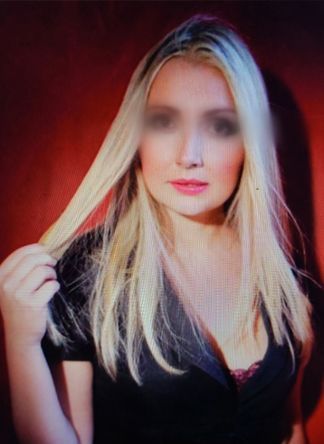 A person with blonde hair stands before a dark red background, wearing a black shirt and holding a strand of their hair.