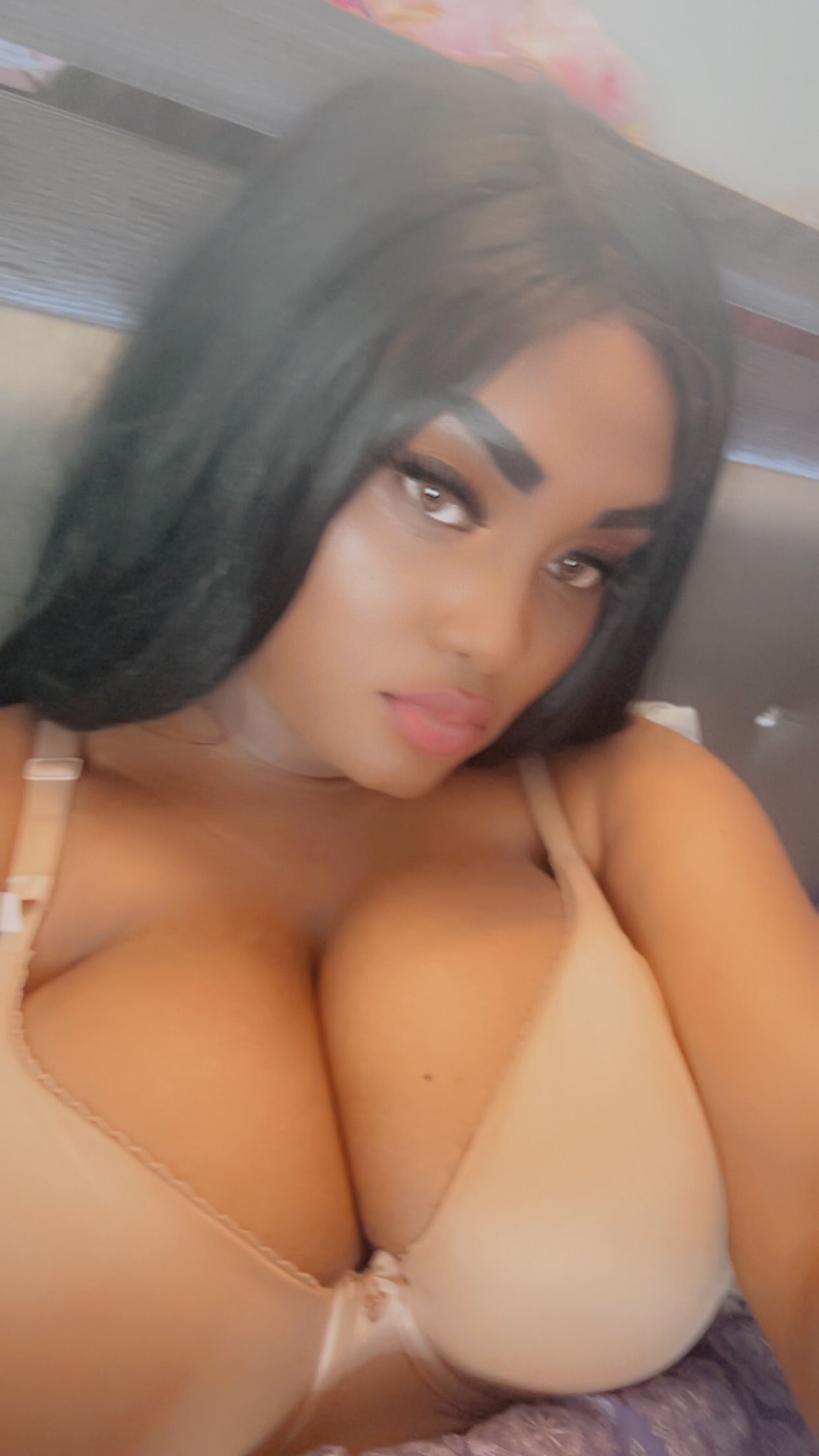 Woman with long black hair and full bust wearing a beige bra, looking at the camera.