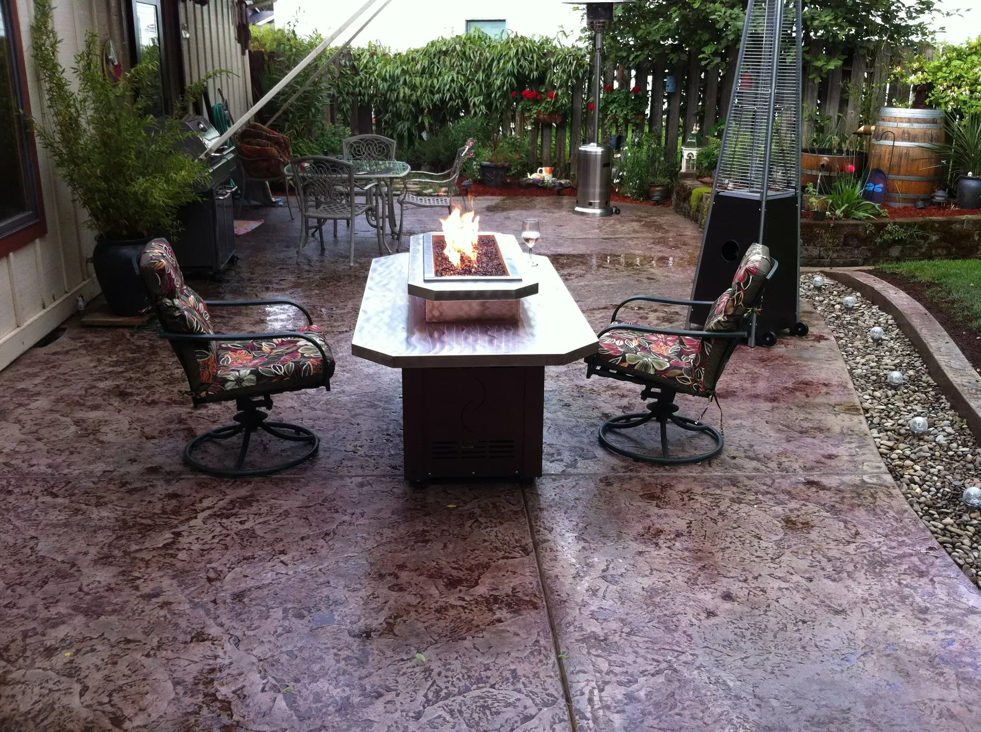 Patio with lit fire pit, two chairs, and stamped concrete flooring. Lush greenery and outdoor heater in background.