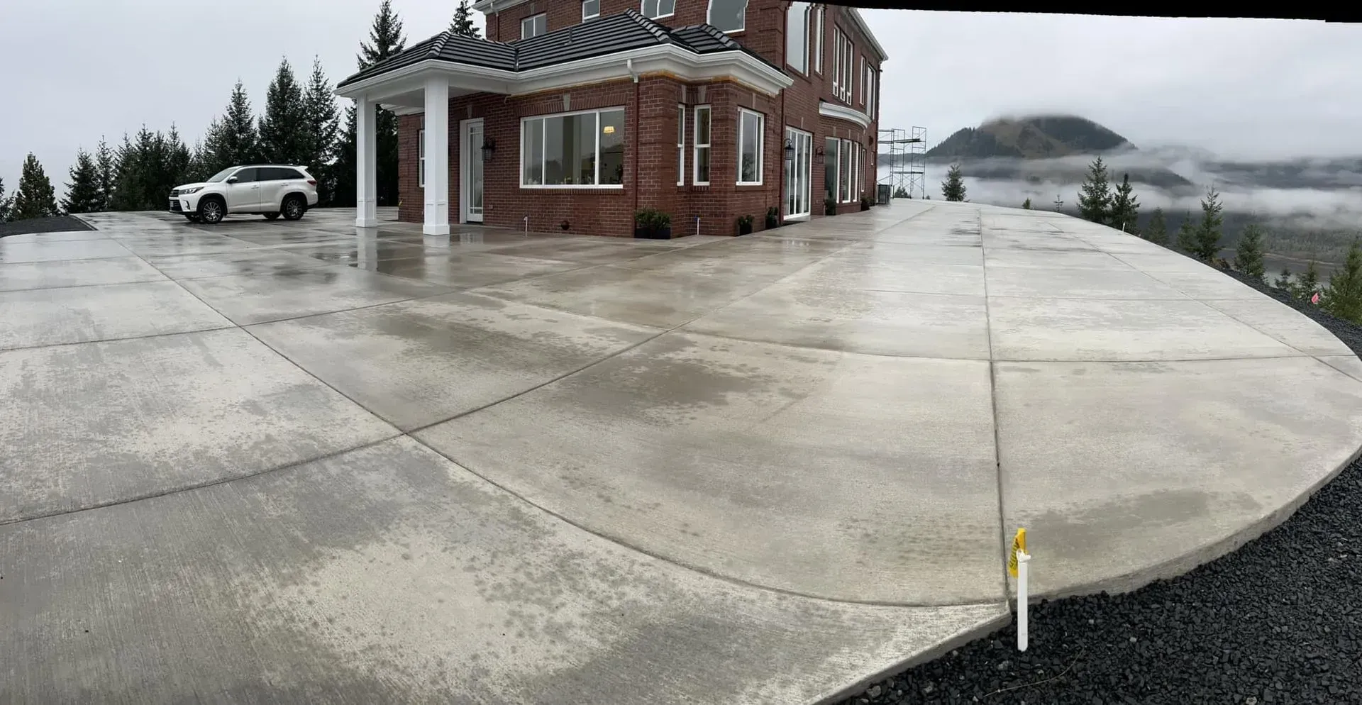 Wet concrete driveway leading to a brick house with a car parked in front. Overcast day.
