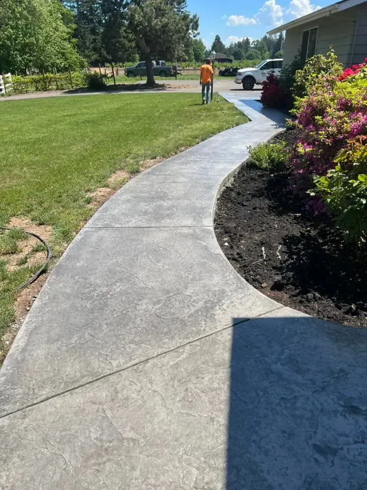 Curving concrete walkway leading to a house, edged with a dark mulch bed and green lawn.
