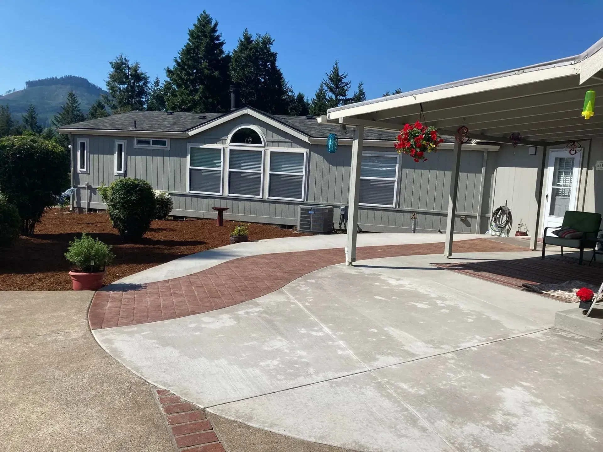 Gray mobile home with brick walkway, covered patio, trees, and mountain backdrop.