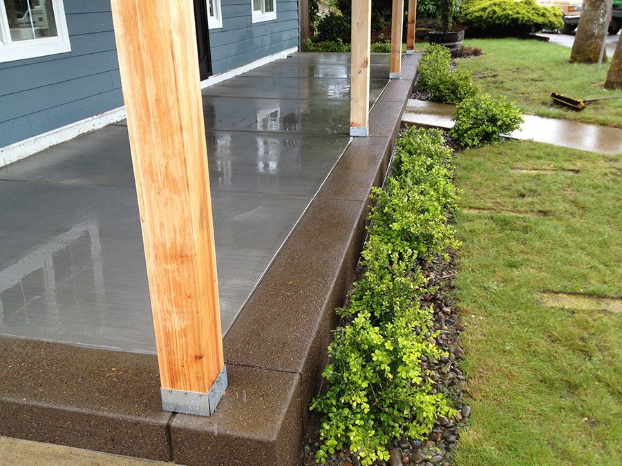 A newly constructed concrete porch with wood support beams and a row of green bushes along the edge.