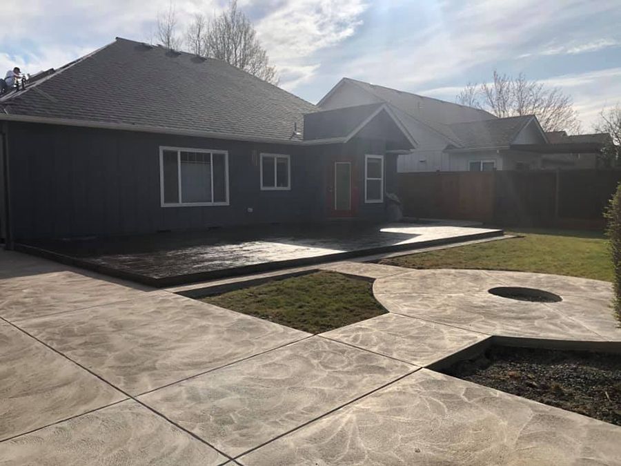 Backyard with gray house, patio, and lawn. Concrete walkway with decorative patterns, and fire pit area.