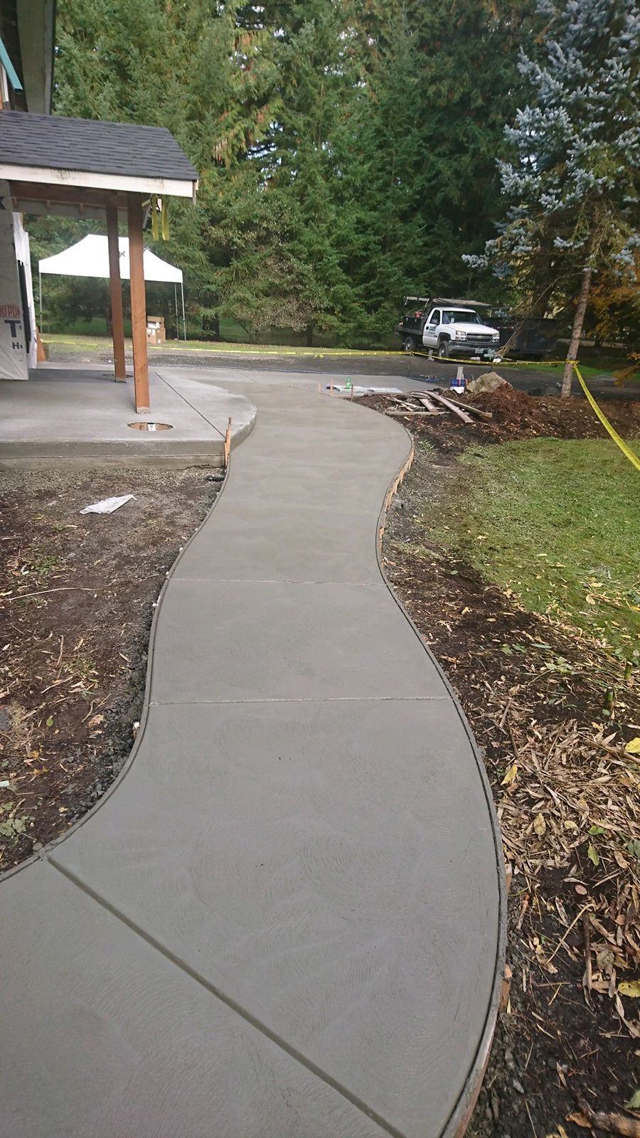 Newly poured concrete walkway curves from a house, through a yard with trees, to a vehicle.