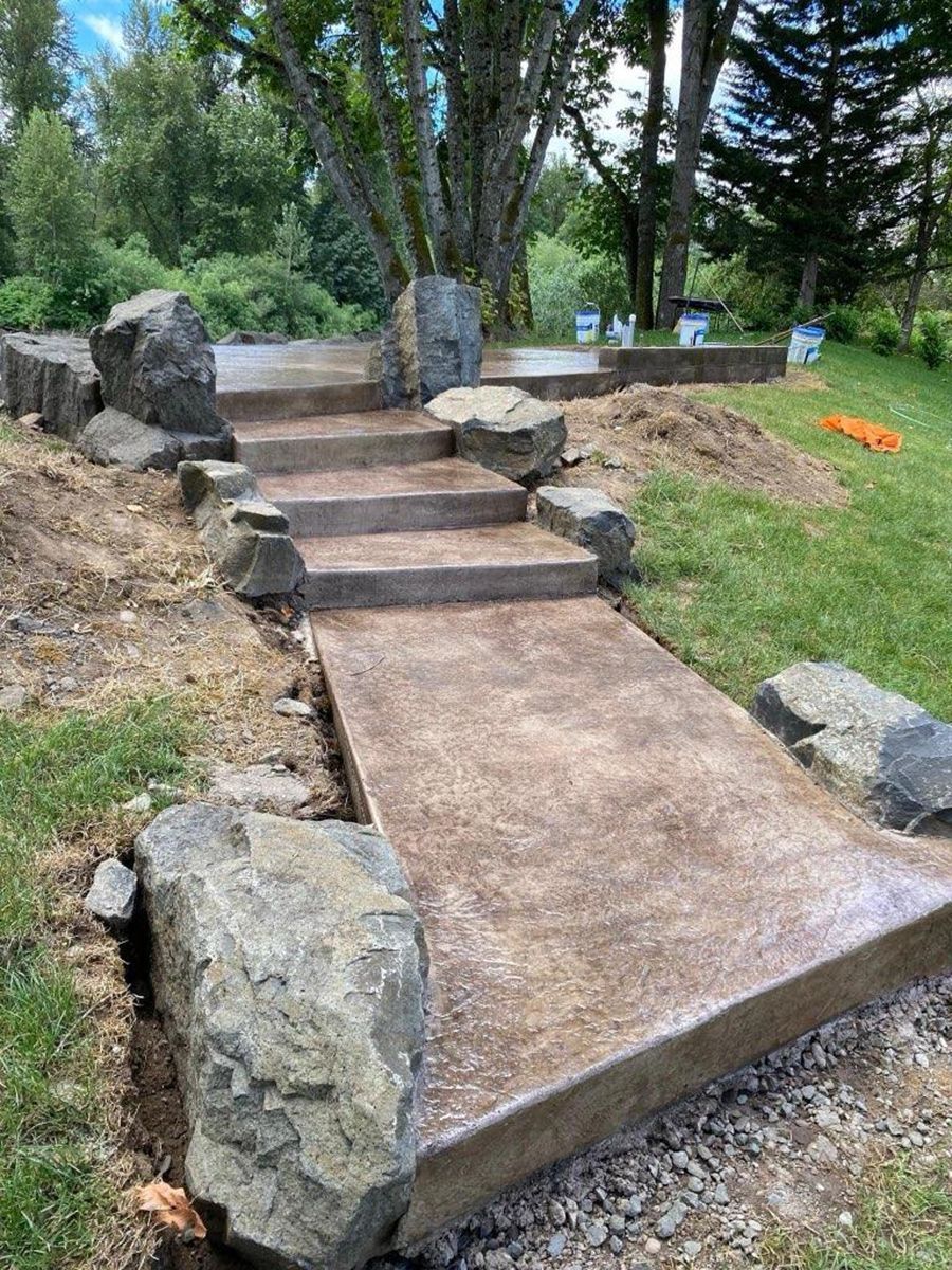 Concrete walkway and steps with large rock borders, leading uphill through a grassy area.