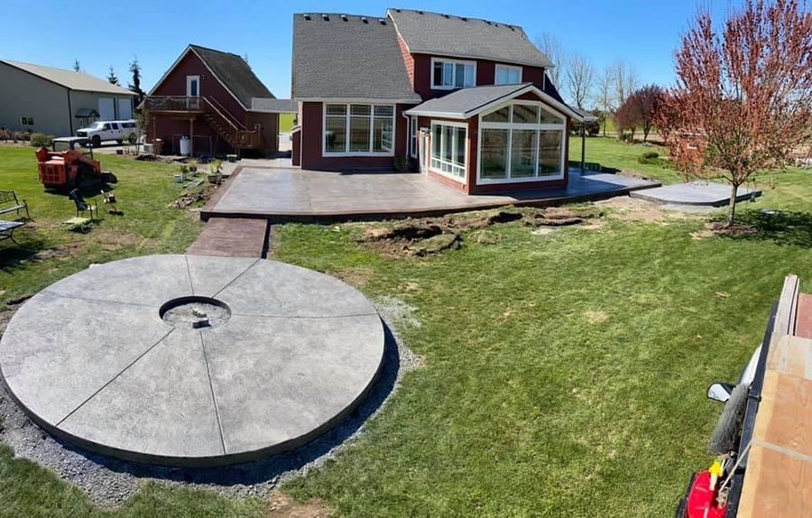 Backyard with house, patio, and large circular fire pit. Reddish-brown buildings, green grass, and blue sky.