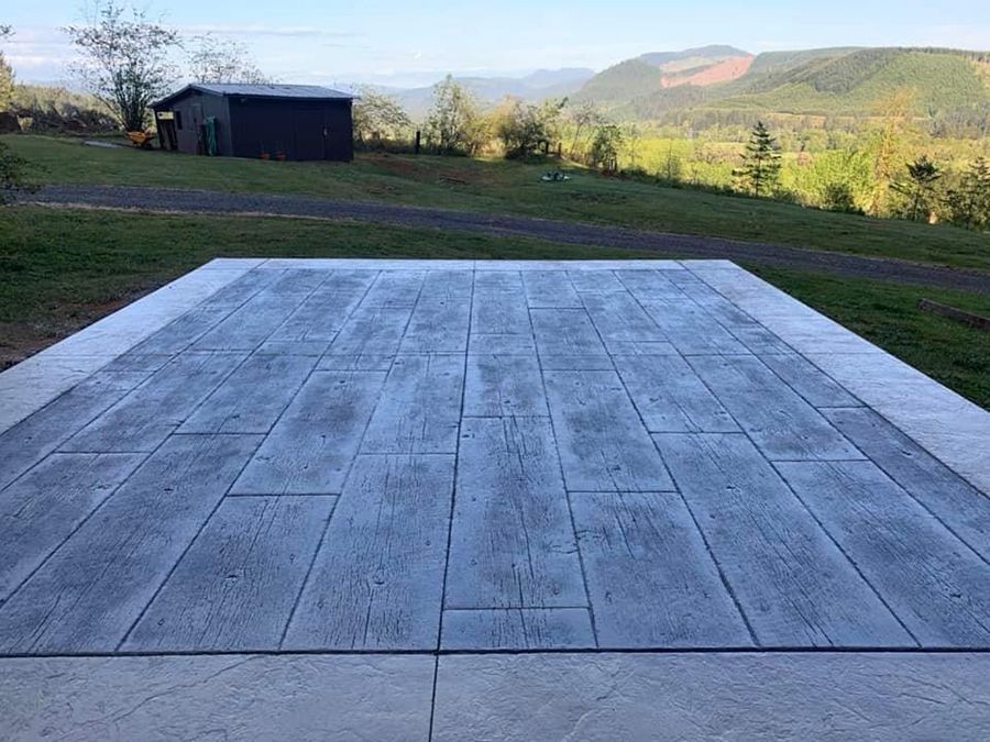 Concrete patio with wood-plank pattern. Green field with distant trees and mountains. Dark shed in the background.