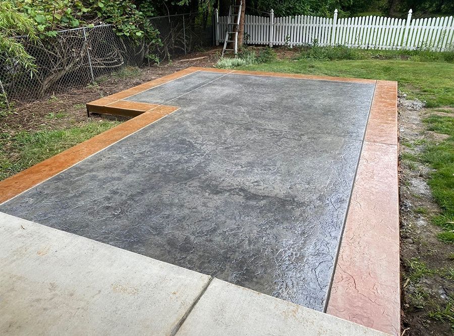 Concrete patio with a brown border and grey textured center, surrounded by grass and fencing.