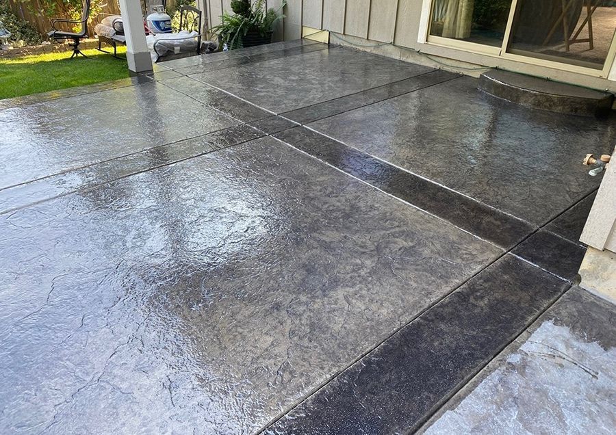 Wet, stamped concrete patio with dark border and lighter center, reflecting sunlight.