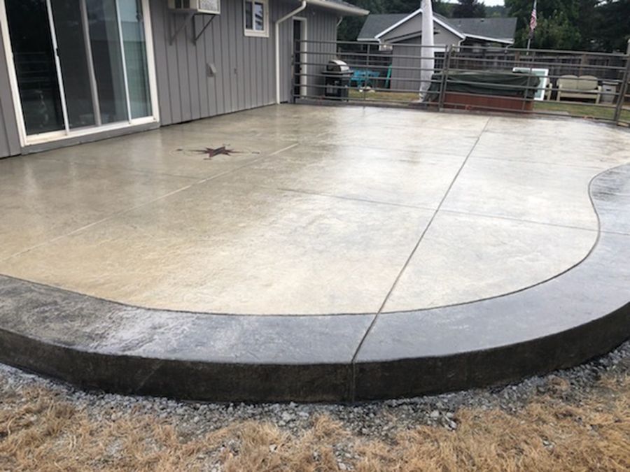 Newly poured concrete patio, light gray with a dark gray curved border, next to a house, outdoor setting.