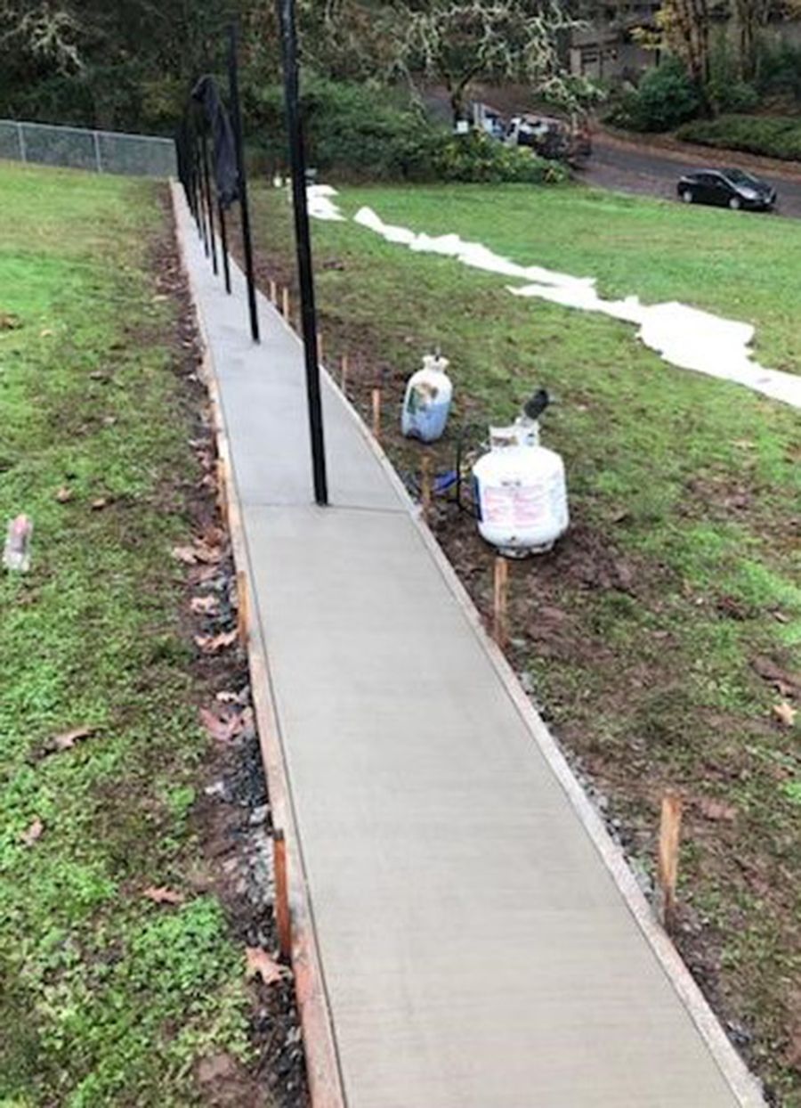 Newly poured concrete sidewalk with black fence, set along a grassy slope, with propane tanks.