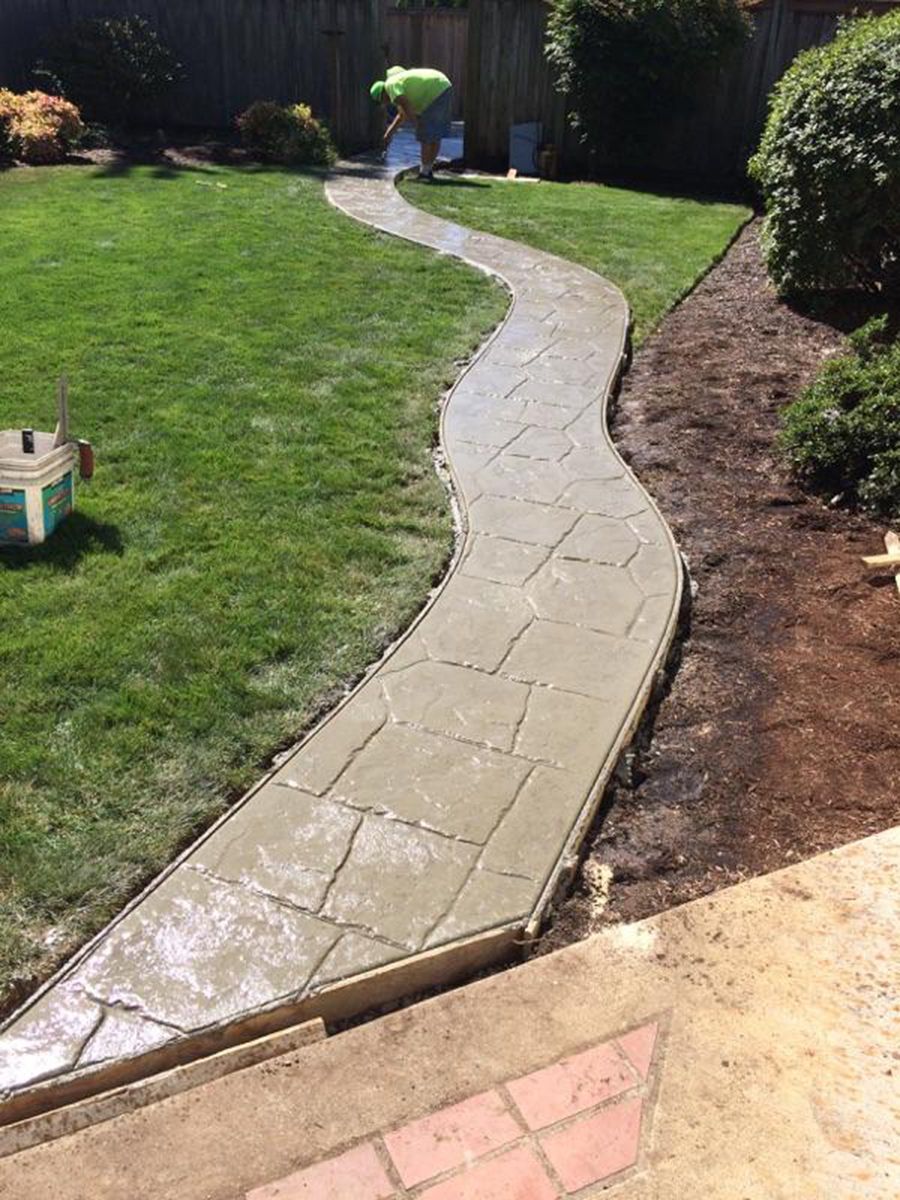 A winding concrete pathway with stone-like texture in a grassy yard. A worker is in the distance.