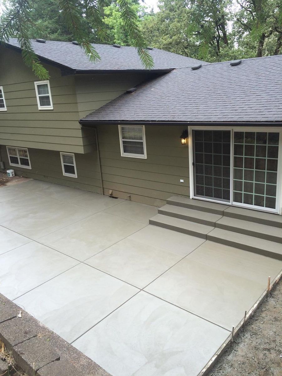 Newly poured concrete patio next to a two-story green house with a sliding glass door and steps.
