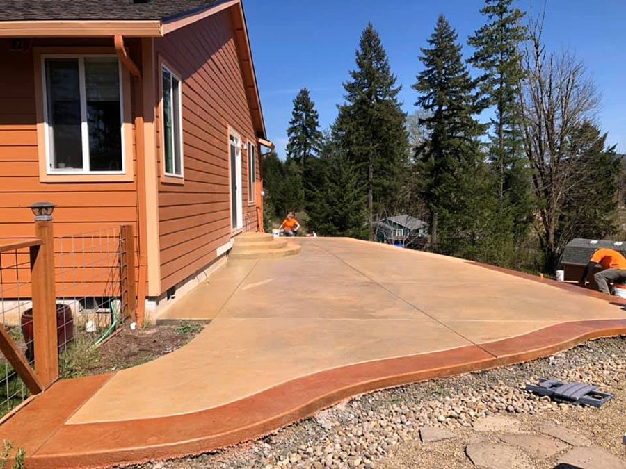 Newly poured concrete patio with reddish-brown border next to an orange-sided house; two workers.