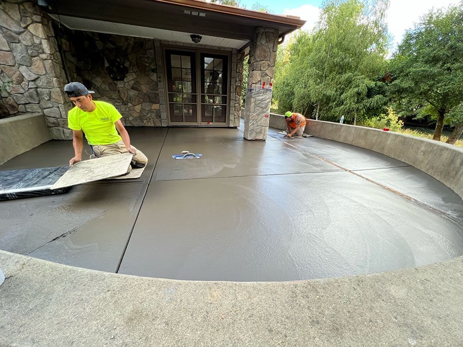 Two workers laying tile on a freshly poured concrete patio in front of a stone-faced building.