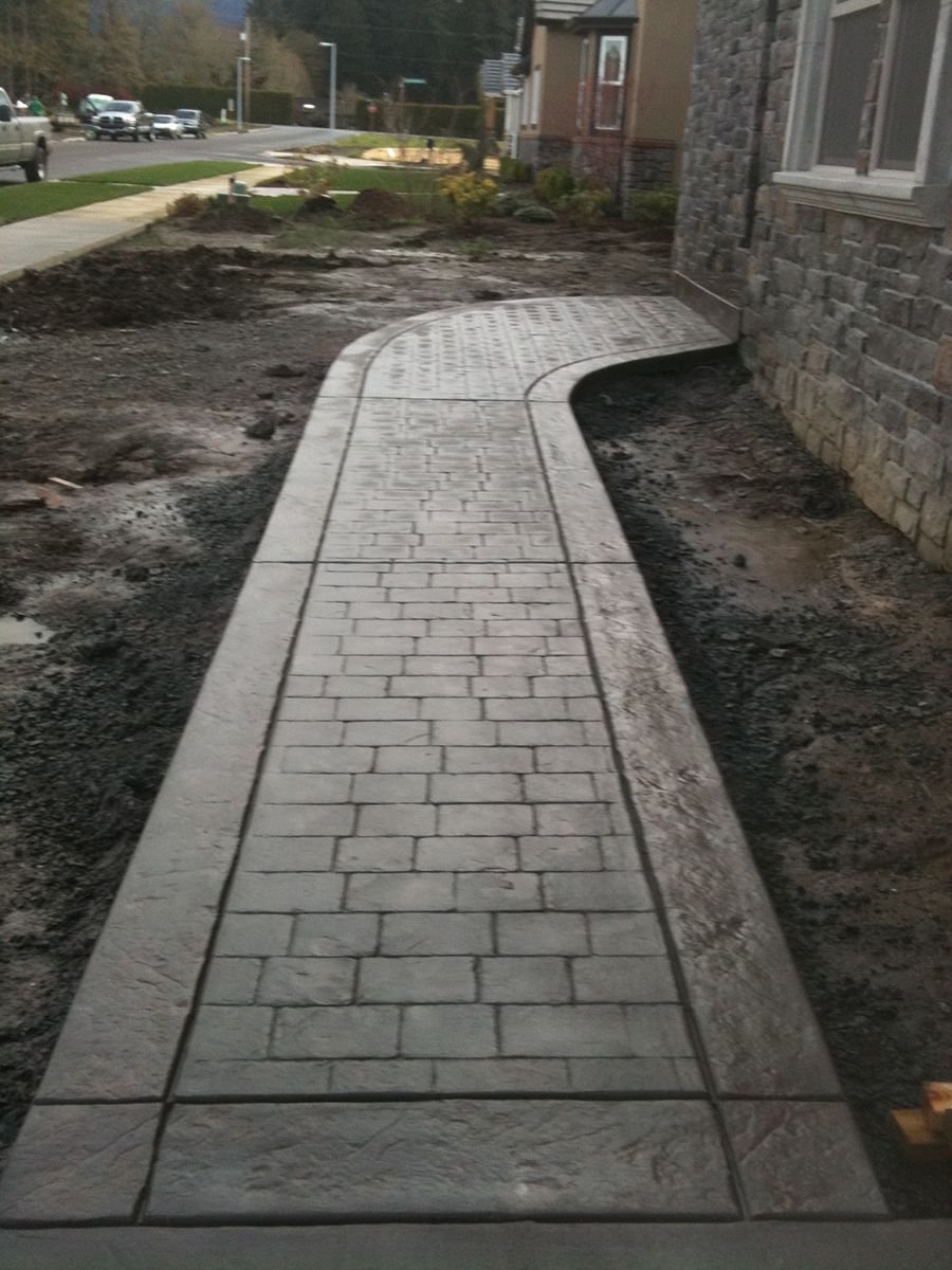 Gray brick-patterned concrete sidewalk curves beside a building, dirt on either side.
