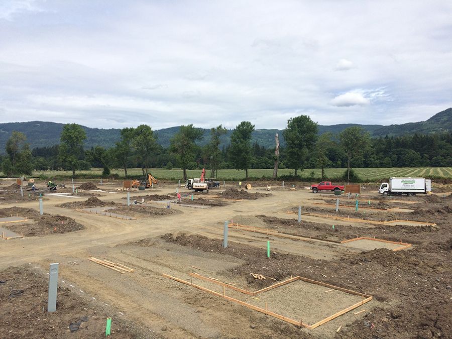 Construction site with earth-moving equipment, trenches, and concrete pillars; mountains in the background under cloudy sky.