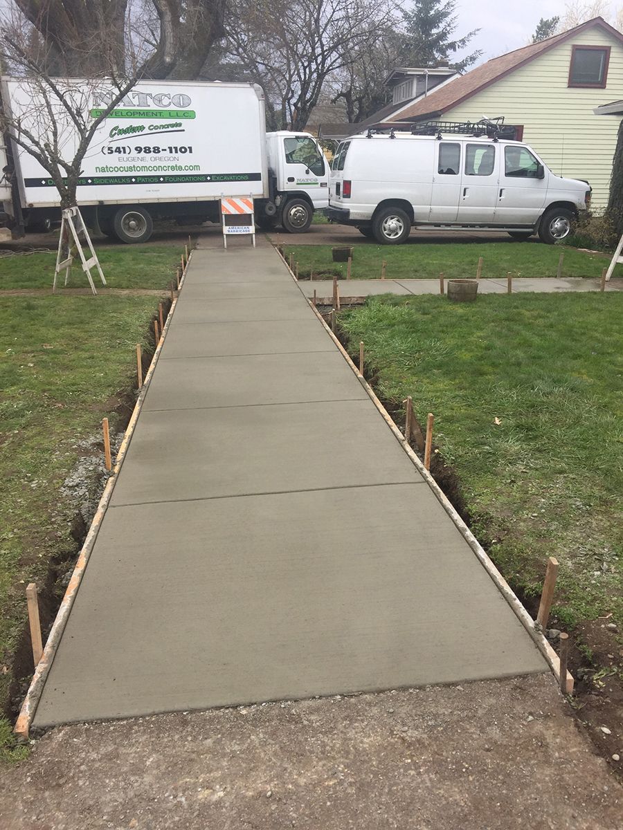 Newly poured concrete sidewalk with wooden formwork, surrounded by grass and parked work vehicles.