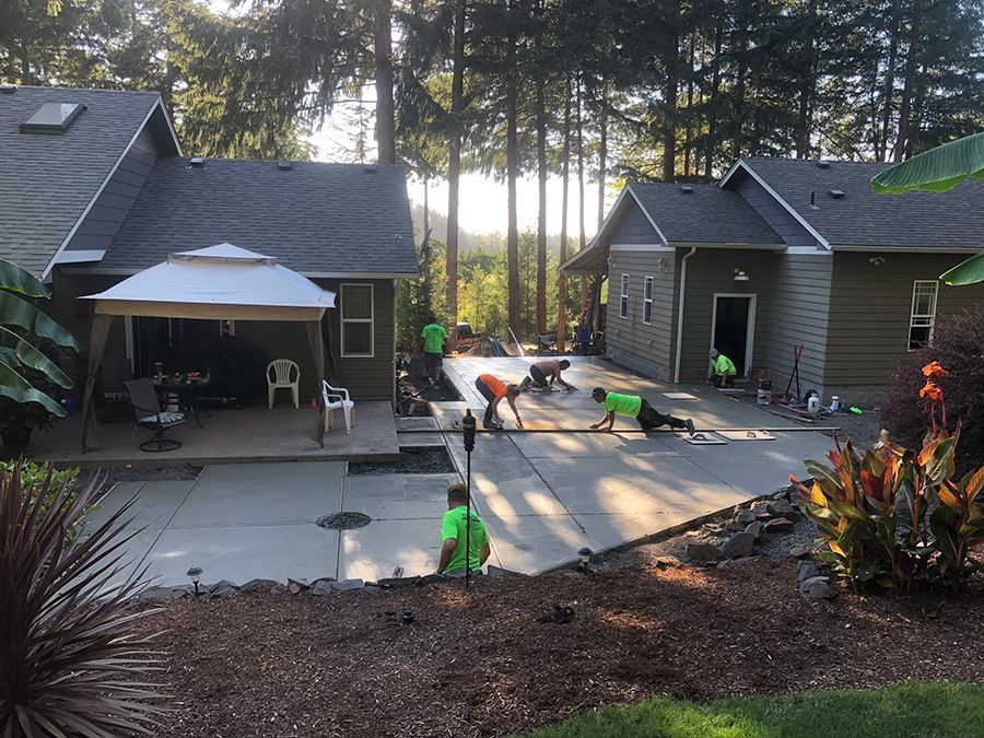 Workers laying concrete patio next to two houses. Sunny day, trees in background, one person standing.