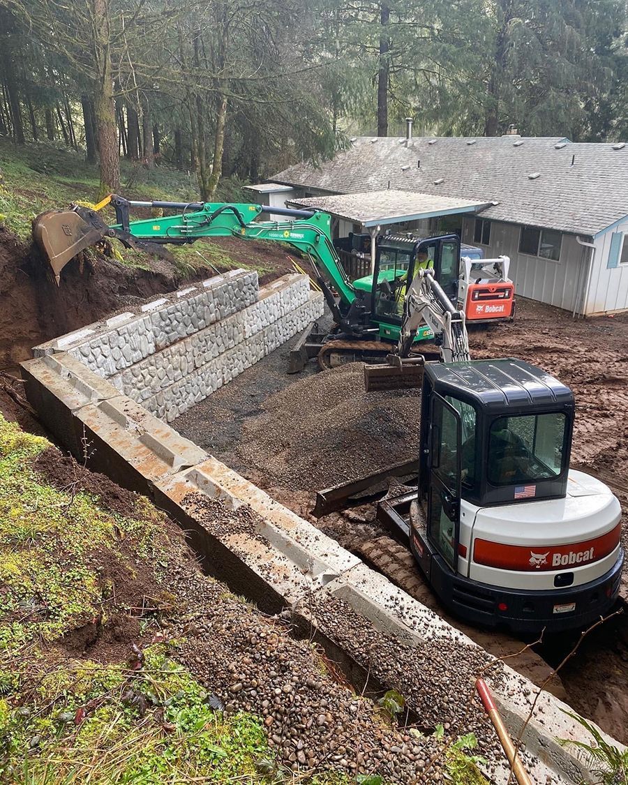 Construction site with excavator and Bobcat, building a retaining wall near a house.