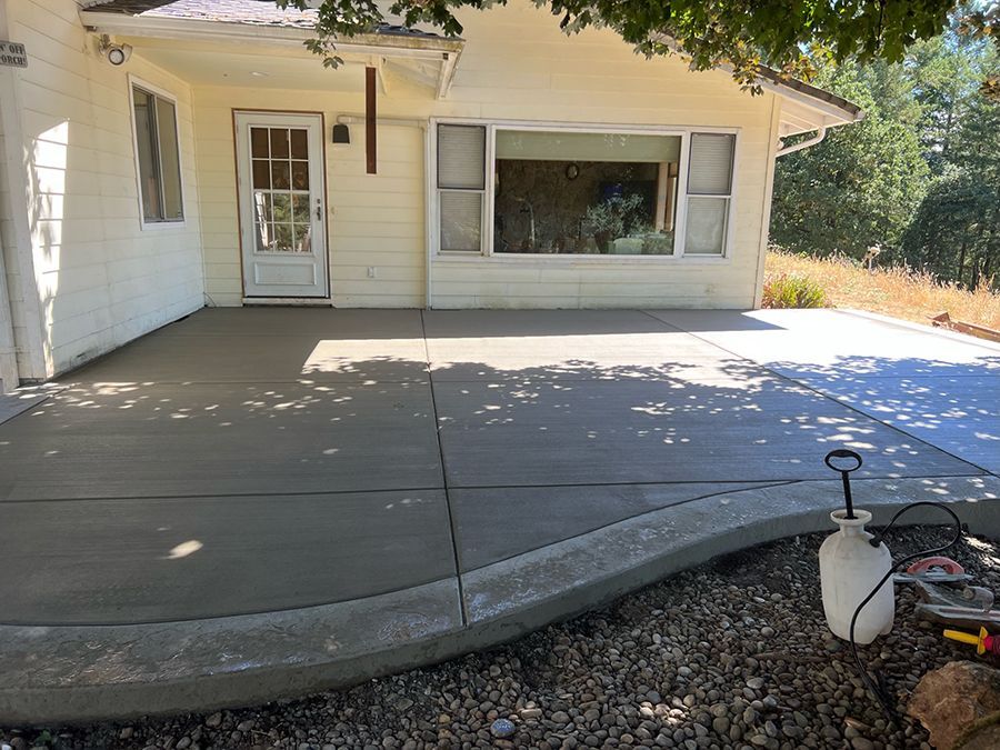 Newly poured concrete patio next to a light yellow house with a door, window, and greenery.