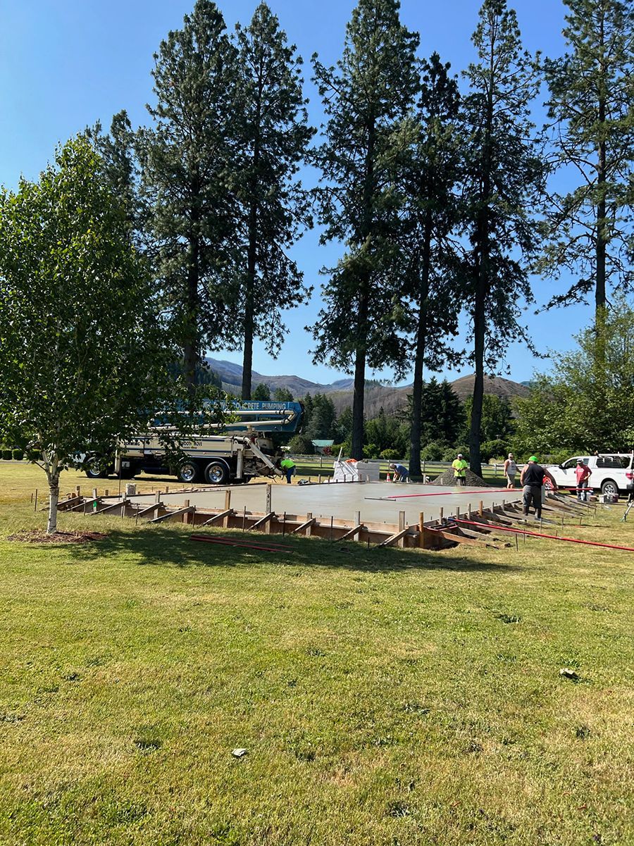 Construction of concrete foundation on a grassy field, trees in the background, blue sky.