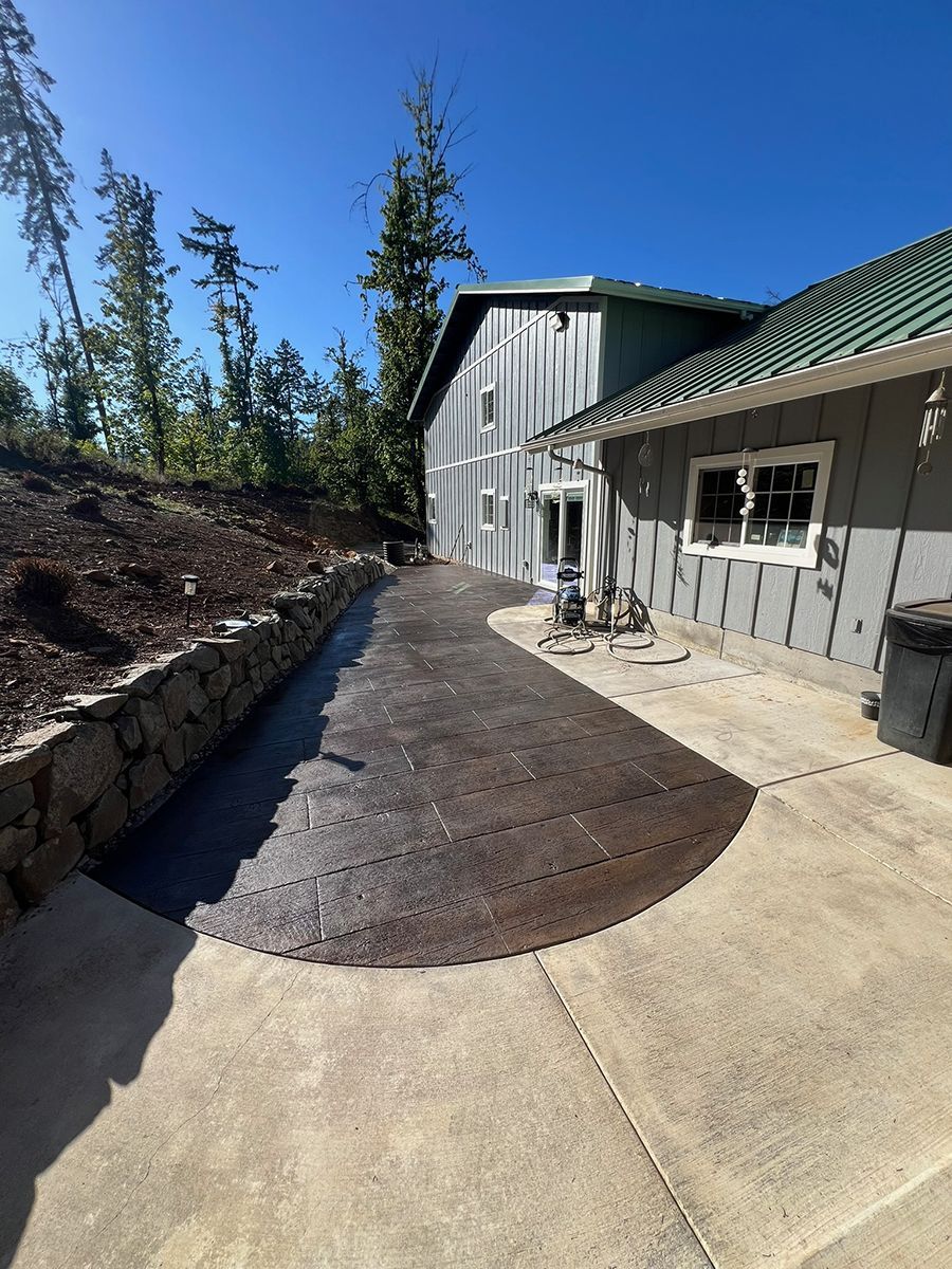 Concrete patio with textured, dark brown section. A building with a green roof is to the right, retaining wall to the left.
