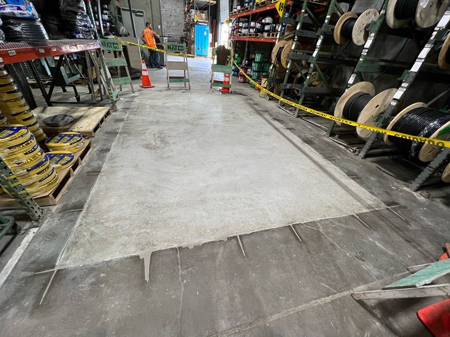 Warehouse floor with fresh concrete patch, caution tape, and electrical cable spools.