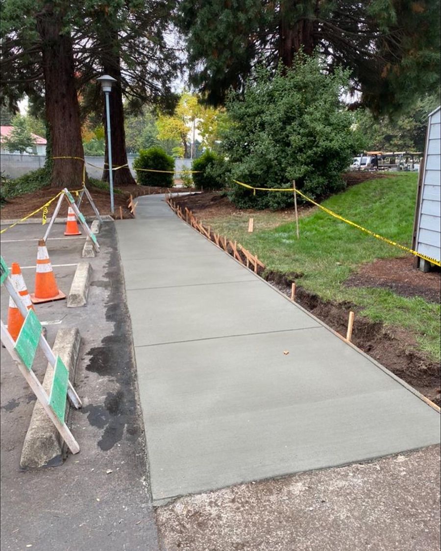 Newly poured concrete sidewalk with caution tape and cones in a park setting.