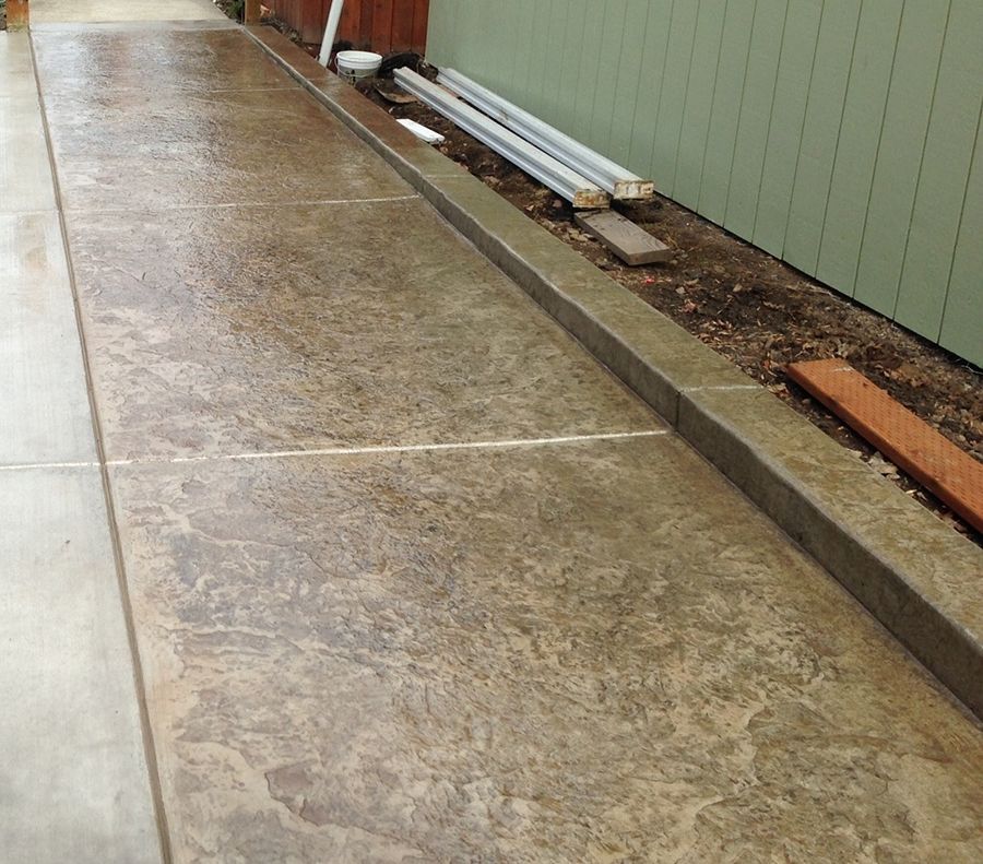 Wet, textured concrete walkway with a raised border next to a green wall and dirt.