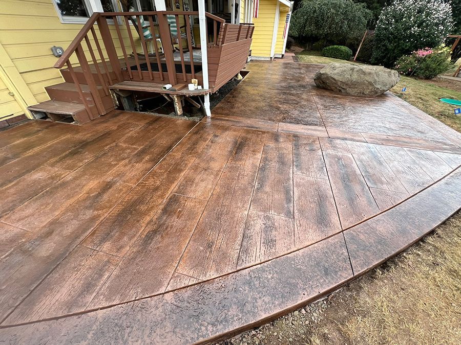 Stamped concrete patio with wood-like texture and brown color, next to a deck and house.