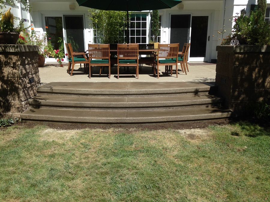 Concrete steps leading to a patio with outdoor dining set; green grass in foreground, white building in background.