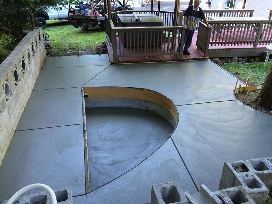 Newly poured concrete patio with a curved cutout and a person standing near a wooden deck.